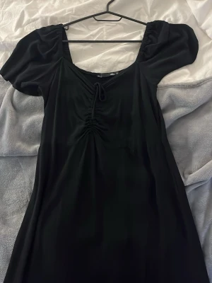 Svart klänning med snörning - Snygg svart klänning från Vila. Framtill finns en dekorativ snörning som ger en fin detalj. Klänningen har en mjuk, följsam passform och är perfekt för dig som gillar enkel men trendig stil.