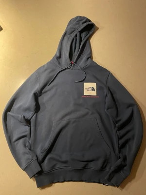 Mörkblå hoodie från The North Face - Snygg mörkblå hoodie från The North Face med stor huva och känguruficka. På bröstet finns en beige patch med loggan och texten 'Never Stop Exploring' i rött. Tillverkad i mjukt bomullsmaterial och har ribbade muddar vid ärmar och nederkant.