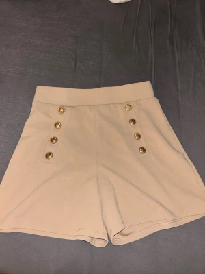 Beige shorts med guldknappar - Stilrena beige shorts med hög midja och sex dekorativa guldknappar framtill. Klassisk och trendig design som passar perfekt till sommaren. Tillverkade i ett mjukt och bekvämt material med lätt stretch för extra komfort.