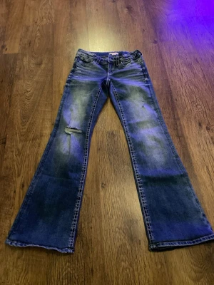 Blå bootcut jeans med broderade fickor - Säljer ett par blå bootcut jeans med slitningar och en liten reva på ena benet. Jeansen har snygga broderade bakfickor med knappar och kontrastsömmar. Perfekt för dig som gillar en avslappnad men ändå trendig stil.