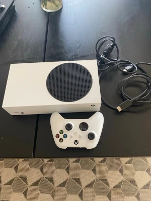 Xbox Series S med handkontroll - Säljer en vit Xbox Series S med tillhörande vit trådlös handkontroll och strömkabel. Konsolen har en kompakt, rektangulär design med en stor svart cirkulär ventil på ovansidan. Perfekt för gaming och digitala spel. Pris går att diskutera 
