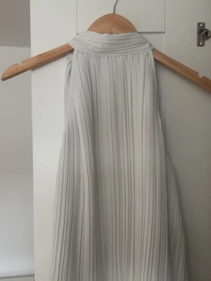 Topp från Zara  - Säljer en stilren ärmlös blus i ljusgrått med plisserat tyg och hög krage. Blusen har en luftig och flowy passform, perfekt för att skapa en chic och modern look. Materialet känns lätt och svalt, vilket gör den skön att bära under varma dagar.