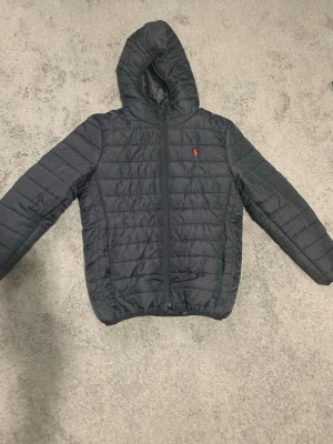 Svart pufferjacka från Polo Ralph Lauren - Snygg svart pufferjacka från Polo Ralph Lauren med huva och dragkedja framtill. Jackan har det klassiska röda Polo-logotypen broderad på bröstet och är quiltad för extra värme. Perfekt för kalla dagar och riktigt stilren look. Priset kan diskuteras vid snabb affär.🔥🔥