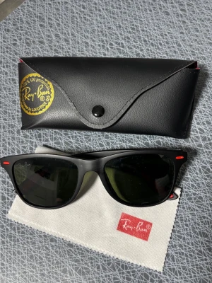 Ray-Ban svarta solglasögon med röda detaljer - Snygga svarta Ray-Ban solglasögon med röda detaljer på skalmarna. Kommer med originalfodral i svart läder och putsduk med Ray-Ban-logga. Klassisk rektangulär form som passar de flesta stilar.