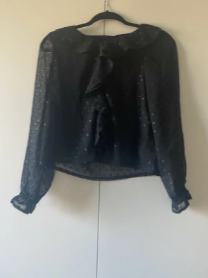 Svart transparent blus med volang - Snygg svart blus i transparent material med små prickar och volangdetalj framtill. Blusen har långa ärmar med resår vid ärmslut och en lös passform. Perfekt för dig som gillar trendiga detaljer och en luftig känsla.