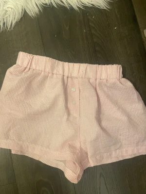 Rosa pyjamasshorts från SHEIN - Söta rosa pyjamasshorts från SHEIN med vita ränder och två små knappar framtill. Shortsen har resår i midjan och är gjorda i ett mjukt, tunt tyg som känns luftigt och bekvämt. Perfekta för mysiga hemmakvällar eller sommarens varma nätter.