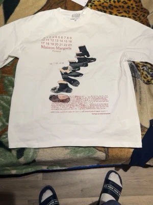 Vit Maison Margiela t-shirt med tryck - Säljer en vit t-shirt från Maison Margiela med unikt grafiskt tryck framtill och logotyp. T-shirten har rund hals och korta ärmar, perfekt för dig som gillar streetwear och designerplagg. Materialet känns mjukt och skönt, troligtvis bomull.