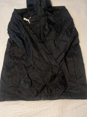 Svart vindjacka från Puma - Snygg svart vindjacka från Puma med dragkedja framtill och huva. Jackan har två sidofickor och elastiska muddar vid ärmsluten. Tillverkad i lätt och tåligt syntetmaterial, perfekt för blåsiga dagar. Puma-logga i vitt på bröstet ger en sportig look. Finns inbyggd luva 