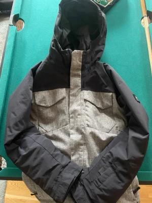 Svart och grå vind/skidjacka från Burton - Snygg skid/vindjacka från Burton i svart och grått med huva och dragkedja. Jackan har två bröstfickor med lock, justerbara ärmslut och en skön innerlining. Perfekt för blåsiga dagar och outdoor-aktiviteter. Stilsäker design med diskret logga på ärmen.