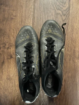 Nike Mercurial svarta fotbollsskor - Säljer ett par Nike Mercurial fotbollsskor i svart med guldiga detaljer och vita inslag. Skorna har snörning och är tillverkade i syntetmaterial för bra grepp och kontroll på planen. Snygg, sportig design som sticker ut på fotbollsplanen.