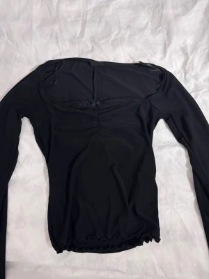 Svart långärmad topp från Gina Tricot - Trendig svart långärmad topp från Gina Tricot med rynkdetalj framtill och liten rosett vid urringningen. Toppen har en figurnära passform och vågig kant nertill. Perfekt för att lyfta din outfit med en enkel men snygg detalj.