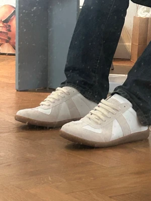 Maison Margiela Gats - Snygga vita sneakers från Maison Margiela med beige mockadetaljer och klassisk gummisula. Skorna har en stilren design med vita skosnören och paneler i både läder och mocka. Perfekta för dig som gillar minimalistisk och trendig stil.