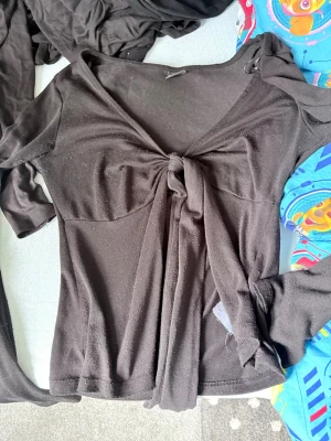 Svart knytblus från Gina Tricot - svart långärmad topp från Gina Tricot med snygg knytning framtill. Toppen har en djup v-ringning och är tillverkad i stretchigt material som sitter bekvämt på kroppen