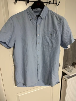 Ljusblå skjorta Dressmann - Kortärmad skjorta från Dressmann i ljusblå bomull. Klassisk button-down krage och bröstficka med knapp. Perfekt till sommaren och enkel att matcha med jeans eller shorts.