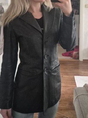 Svart kappa/kavaj i äkta skinn - Snygg svart kappa/kavaj i äkta skinn med figurnära passform och klassisk krage. Kappan har tre knappar framtill och två fickor på sidorna. Perfekt för dig som vill ha en stilren och edgy look.