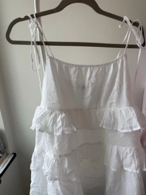 Vit volangklänning med knytband - Söt vit klänning med tunna axelband som knyts på axlarna och flera volanglager. Tillverkad i ett lätt, krinklat tyg som ger en luftig känsla. Perfekt för varma dagar och har en avslappnad, flowy passform.