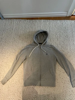 Grå hoodie från Polo Ralph Lauren - Snygg grå zip-hoodie från Polo Ralph Lauren som nästan aldrig är använd med huva och vita dragsnören. Klassisk liten logga broderad på bröstet. Tillverkad i mjukt bomullsmaterial och har två fickor framtill. Perfekt för en avslappnad och stilren look.