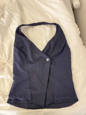 Mörkblå halterneck topp - Snygg mörkblå halterneck topp med omlott-design och knappdetalj framtill. Toppen har en djup v-ringning och är ärmlös, vilket ger en trendig och elegant look. Perfekt att styla med jeans eller kjol för en cool outfit.