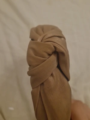 Beige twistat diadem i tyg - Snyggt beige diadem med twistad knut framtill. Tillverkat i mjukt tyg med en matt finish och svart insida för extra komfort. Perfekt accessoar för att lyfta din look med en trendig touch.