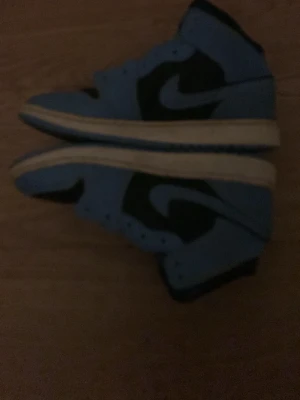 Nike Air Jordan 1 High blå/svart - Säljer ett par Nike Air Jordan 1 High sneakers i blått och svart med vit sula. Klassisk high-top siluett med ikoniska swoosh-loggan på sidan och Jumpman-logga på plösen. Materialet är en mix av läder och textil för en clean och sportig look.