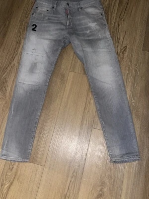 Grå jeans med tryck från Dsquared2 - Säljer ett par grå jeans från Dsquared2 med svart siffra '2' tryckt på vänster lår. Jeansen har en avslappnad passform, klassiska fem fickor och diskreta slitningar för en cool look. Perfekta för dig som gillar streetwear och unik stil.