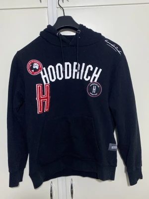 Svart hoodie från Hoodrich med tryck    Den passar M o S - Snygg svart hoodie från Hoodrich med vita och röda tryck och broderade detaljer på bröstet. Stor luva med dragsko, känguruficka framtill och ribbade muddar. Hoodrich-logga på bröstet och patchar för en streetwear-look. Perfekt för dig som gillar urban stil.