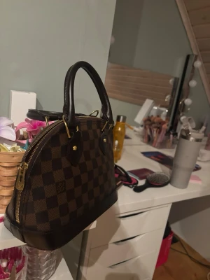  brun rutig handväska - Säljer en ikonisk handväska  i klassiskt brunt rutigt mönster med guldfärgade detaljer och dragkedja. Väskan har två handtag, rundad form och röd insida. Tillverkad i skinn och canvas, perfekt för dig som gillar lyxiga accessoarer. Band följer med pris går att diskutera vid snabbköp (lås finns ej)