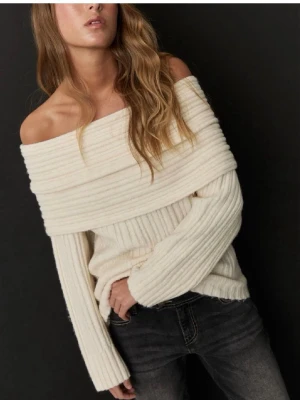 Off shoulder stickad tröja cremevit - Trendig off shoulder stickad tröja i cremevitt med bred ribbstickning. Tröjan har långa ärmar och en bred, nedvikt krage som ger en snygg och avslappnad look. Perfekt för dig som vill ha en stilren och mysig outfit.