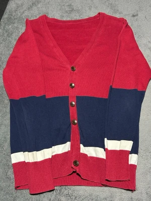 Vintage Style Colour Block Cardigan – Red/Navy – Size M/L - Stickad cardigan i retro colour block design med rött, navy och vita detaljer.  Klassisk modell med knappar framtill och mjukt stickat material. Perfekt för en casual eller vintage inspirerad stil.  Detaljer • Stickad cardigan • Colour block design • Röd / navy / vit • Knappar framtill • Mjuk stickning  📦 Skickas snabbt 💬 Hör av dig vid fråga