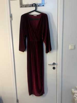 Vinröd långklänning i sammet - Elegant vinröd långklänning i mjuk sammet med v-ringad omlottfront och långa ärmar. Perfekt för dig som vill ha en lyxig och stilren look. Klänningen har markerad midja och faller snyggt längs kroppen.