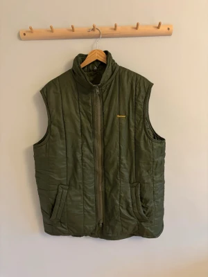 Olivgrön dunväst från Barbour - Snygg olivgrön dunväst från Barbour med gul broderad logga på bröstet. Västen har hög krage, dragkedja framtill och två fickor. Perfekt för lager-på-lager och ger en stilren look. Tillverkad i quiltat material som håller värmen.