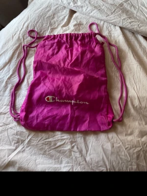 Rosa gympåse från Champion - Säljer en snygg rosa gympåse från Champion med dragskostängning och tunna axelband. Väskan har ett litet blixtlåsfack på baksidan och Champion-loggan tryckt på framsidan. Perfekt för träning eller skola, enkel att bära med sig. Materialet är lätt och smidigt.