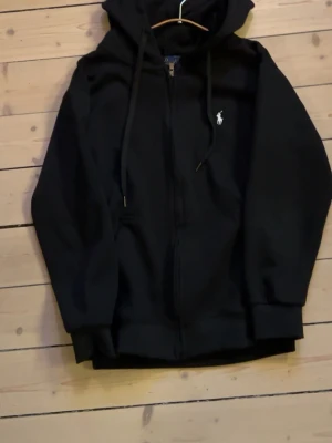 Svart hoodie från Polo Ralph Lauren - Säljer en svart hoodie från Polo Ralph Lauren med dragkedja framtill och klassisk vit logga på bröstet. Hoodien har justerbar huva med snören och ribbade muddar. Perfekt för en chill och stilren look. Endast använd 1 gång. Skriv dm för mer bilder/ frågor. MVH John💛