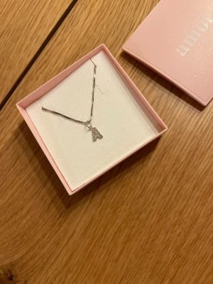 Silverhalsband med bokstav A från Amor - Snyggt silverfärgat halsband från Amor med ett hänge i form av bokstaven A, dekorerat med små glittrande stenar. Kedjan är tunn och elegant, perfekt för att ge en personlig touch till din stil. Levereras i en söt rosa ask.