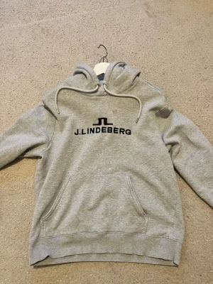 Grå hoodie från J.Lindeberg - Snygg grå hoodie från J.Lindeberg med svart logga på bröstet. Klassisk modell med huva, dragsnören och stor magficka. Mjuk bomullsblandning och ribbade muddar vid ärmslut och nederkant. Perfekt för en chill och stilren look. (Bild 3 litet hål väldigt enkelt att laga tar max 3 min!)