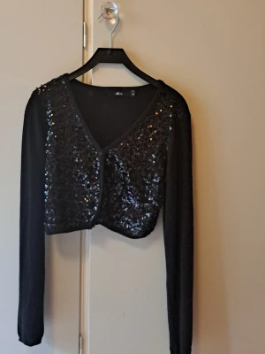 Svart paljettkofta/bolero från Ellos - Detta är en kort croppad kofta / bolero från Ellos. Köpt 2011, finns ej mer. Den har glittriga paljetter framtill. Man kan ha något under eller inget o därmed cropped look. Den är perfekt för nyår, fin fest, klubb, disco, dejt, vinprovning. Den har knappar så man kan ha den både stängd eller öppen. Den är det där perfekta som värmer en på en utekväll utan att vara den där fula koftan över klänning. Tänker på Serena van der woodsen när jag ser den. 