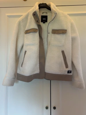 Vit teddyjacka från Vans - Säljer en vit teddyjacka från Vans med beige detaljer. Jackan har två bröstfickor med knappar, två sidofickor och dragkedja framtill. Materialet är mjukt och fluffigt, perfekt för kyliga dagar. Vans-logga på ena fickan och beige kantband längs nederkant och ärmslut.