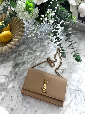 Beige kuvertväska från Saint Laurent - Snygg beige kuvertväska i slätt läder med guldfärgad kedja och ikoniska YSL-loggan framtill. Stilren och elegant design som passar perfekt till alla tillfällen. Väskan har en klassisk rektangulär form och stängs med lock.