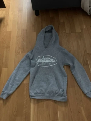 Grå hoodie med skeppstryck - Snygg grå hoodie med ett vitt tryck av ett skepp på bröstet. Hoodien har en stor huva och en klassisk magficka framtill. Perfekt för en chill och avslappnad stil. Materialet känns mjukt och bekvämt, passar bra till jeans eller joggers.