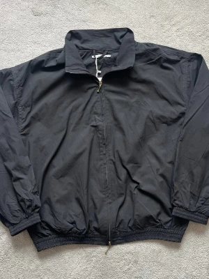 Acne Studios Svart vindjacka med logo baksida - Svart vindjacka med klassisk krage och hel dragkedja framtill. Jackan har resår vid ärmslut och midja för en skön passform. Tillverkad i lätt och vindtätt material, perfekt för blåsiga dagar. Enkel och stilren design utan synliga loggor.