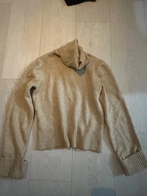Beige stickad polotröja - Mysig beige stickad polotröja med långa ärmar och breda ribbade muddar vid ärmslut och nederkant. Tröjan har en hög, vikt polokrage och är perfekt för kyliga dagar. Materialet ser ut att vara mjukt och värmande.