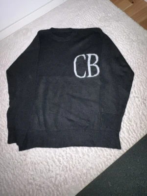 Mörkgrå stickad tröja med CB-logga - Mörkgrå stickad tröja med rund halsringning och vita bokstäver 'CB' broderade på bröstet. Tröjan har långa ärmar och ribbstickade muddar vid ärmslut och nederkant. Perfekt för en stilren och enkel look.