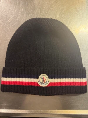  Moncler Tricolor Mössa - Svart ribbstickad mössa från Moncler med uppvikt kant. Mössan har klassiska ränder i blått, vitt och rött samt Moncler-logga framtill. Perfekt accessoar för att hålla värmen och samtidigt se stilren ut.