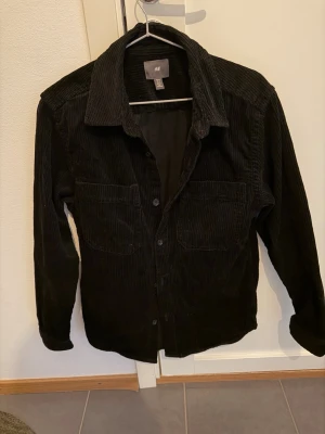Svart manchester-skjorta från H&M - Svart skjorta i manchestertyg från H&M med klassisk krage och två bröstfickor. Skjortan har knäppning framtill och långa ärmar. Perfekt för lager-på-lager och ger en cool vibe till din outfit.