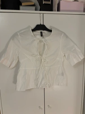 Vit topp med snörning från H&M - Snygg vit topp från H&M med kort ärm och trendig snörning framtill. Modellen är croppad med volang nedtill och har en öppen detalj vid bröstet. 
