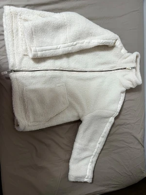 Vit teddyjacka med dragkedja - Mysig vit teddyjacka med hög krage och hel dragkedja framtill. Jackan har två stora fickor på sidorna och är tillverkad i fluffigt teddy-material som ger en cool och avslappnad look. Perfekt för kalla dagar när du vill vara både varm och trendig.