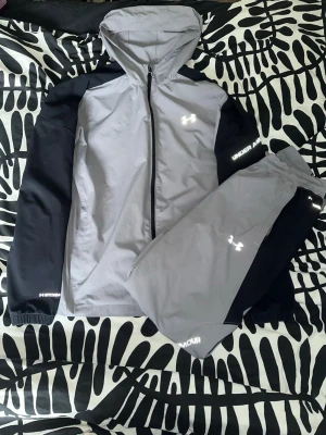 Under Armour grå träningsset - Snyggt träningsset från Under Armour med grå jacka och byxor, svarta detaljer på ärmar och ben. Jackan har huva och dragkedja, byxorna har resår i midjan och dragkedjor vid benslut. Reflekterande loggor och text. Perfekt för löpning eller träning utomhus.