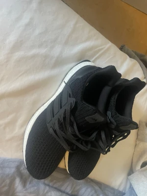 Svarta Adidas Ultraboost sneakers - Svarta Adidas Ultraboost sneakers med stickad ovandel och klassiska tre ränder på sidan. Skorna har en vit Boost-mellansula och svart yttersula med greppvänligt mönster. Perfekta för dig som gillar sportig och stilren look.