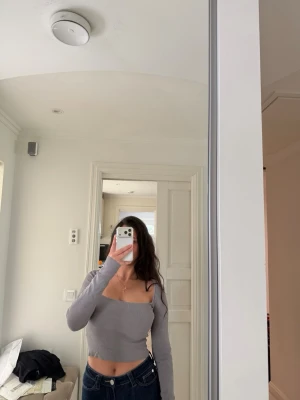 Ljusgrå croppad ribbad topp - Trendig ljusgrå croppad topp med lång ärm och fyrkantig urringning. Toppen är ribbad och har en tight passform som framhäver figuren. Perfekt att styla med höga jeans eller kjol för en cool look.