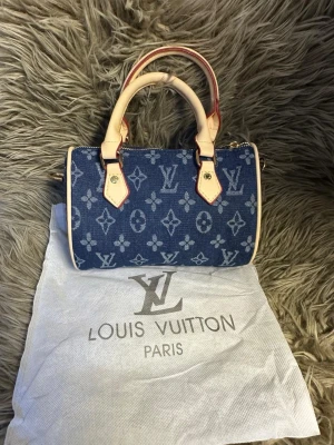  blå monogram handväska - Snygg handväska  i blått denimtyg med klassiskt monogrammönster. Väskan har ljusa skinnhandtag och detaljer, samt guldfärgad dragkedja. Perfekt storlek för det viktigaste och en ikonisk accessoar till din outfit.